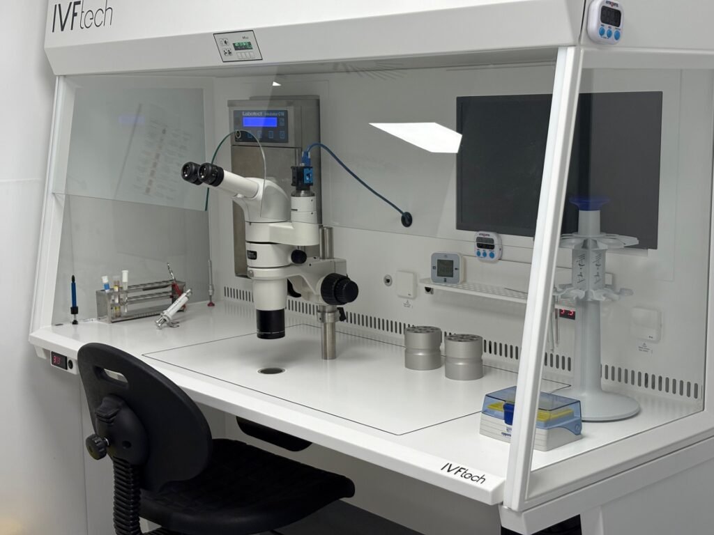 IVF-Arbeitsplatz der Klinik: Sicherheitswerkbank mit Stereomikroskop, Inkubator, Bildschirm und Pipetten hinter einer Glasscheibe.