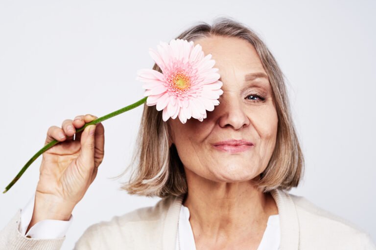 Ältere Frau mit grauem Bob lächelt und hält sich eine rosa Gerbera vor ein Auge.